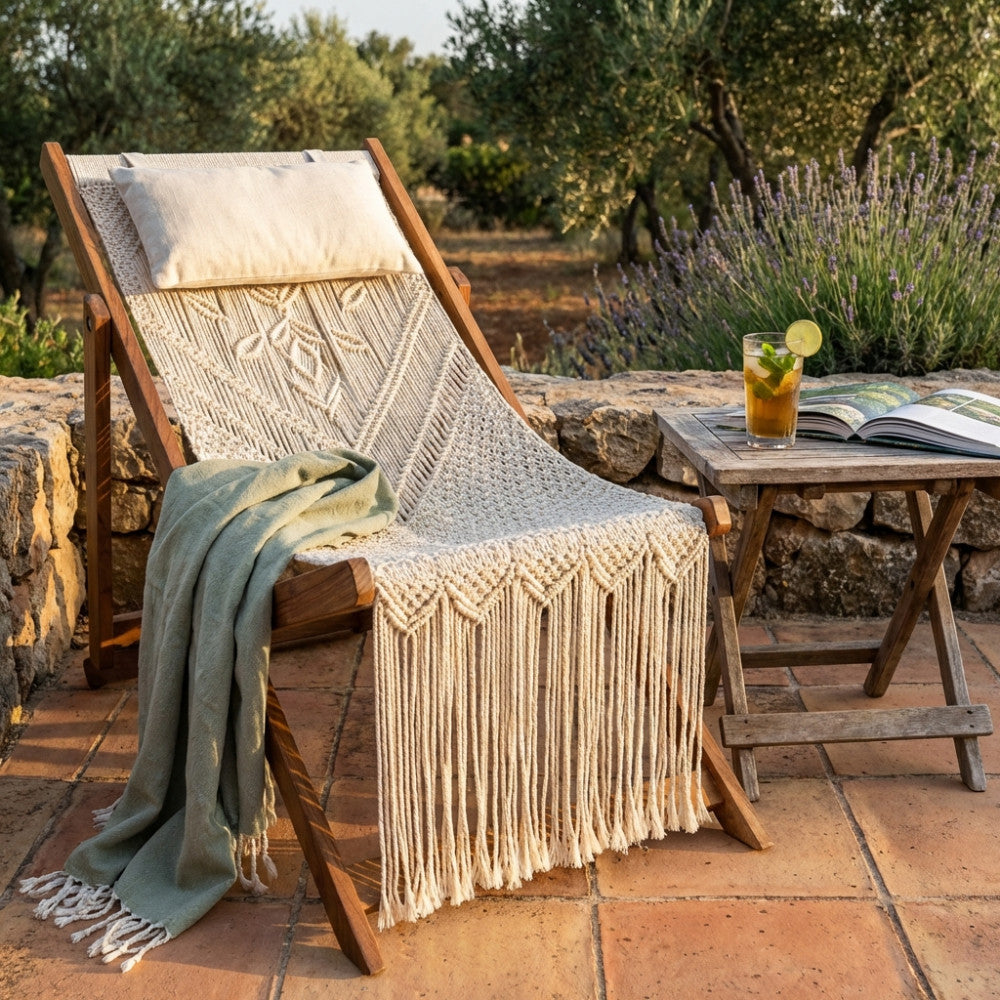 Sedia a sdraio Tunisi in cotone macramè ambientata in un giardino rustico con cuscino, coperta verde e tavolino in legno.
