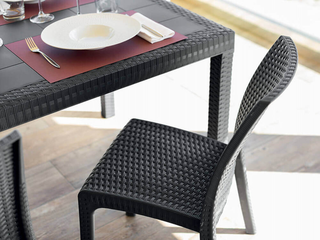 Dettaglio della sedia Virginia in wicker antracite accostata a un tavolo da pranzo scuro finemente apparecchiato.