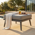 Seduta modulare in alluminio antracite con cuscino grigio chiaro, allestita a bordo piscina con un telo e un vassoio con drink.