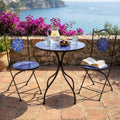 Set bistrot Tiche in ceramica blu con tavolo rotondo e due sedie pieghevoli, allestito su una terrazza in cotto con vista sul mare e bouganville.