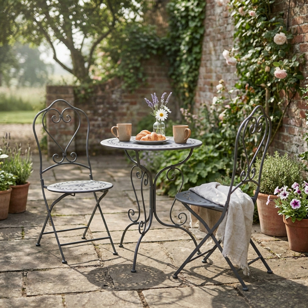 Tavolo e sedie bistrot Jeanette in acciaio decorato, disposti in un giardino fiorito per una colazione all'aperto.