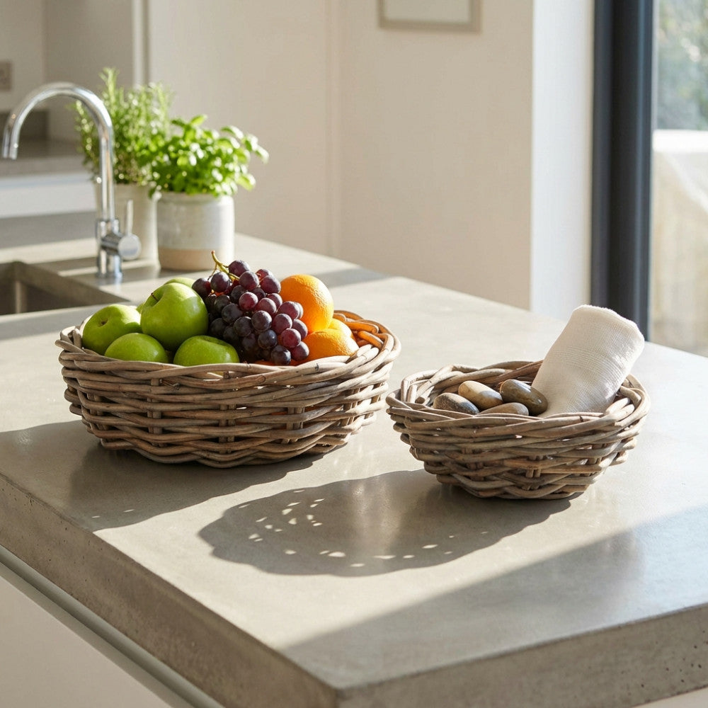 Set di due cesti Serge in vimini grigio posizionati su un piano cucina in cemento, allestiti con frutta fresca e asciugamani in lino sotto la luce naturale.