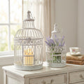 Set di due gabbie decorative in acciaio bianco antico posizionate su un mobile in legno chiaro, adornate con una candela e fiori di lavanda.