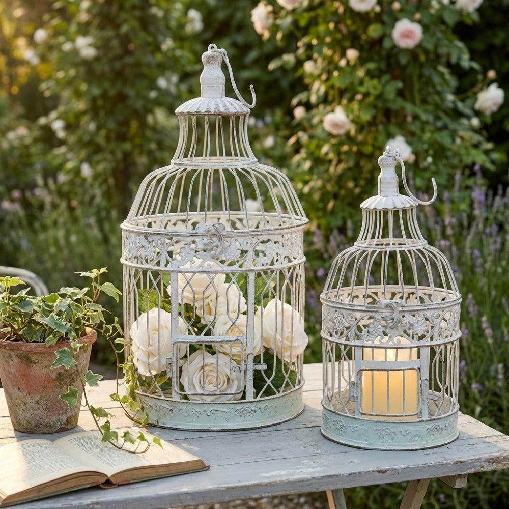 Gabbie decorative bianche in stile provenzale allestite su un tavolo da giardino all'aperto, decorate con rose bianche e una candela accesa.