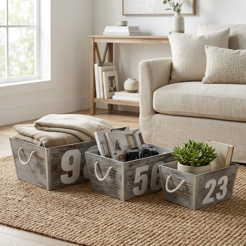 Set di 3 cassette in legno grigio Zoe con numeri decorativi, allestite su un tappeto in iuta con plaid, libri e una pianta in un salotto luminoso.
