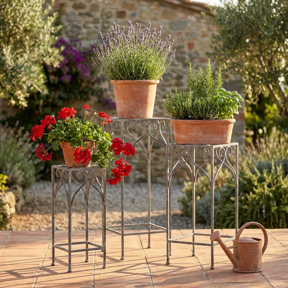 Tre fioriere in acciaio bianco ossidato di diverse altezze in un giardino rustico, arricchite con gerani rossi, lavanda e piante aromatiche in vasi di terracotta.