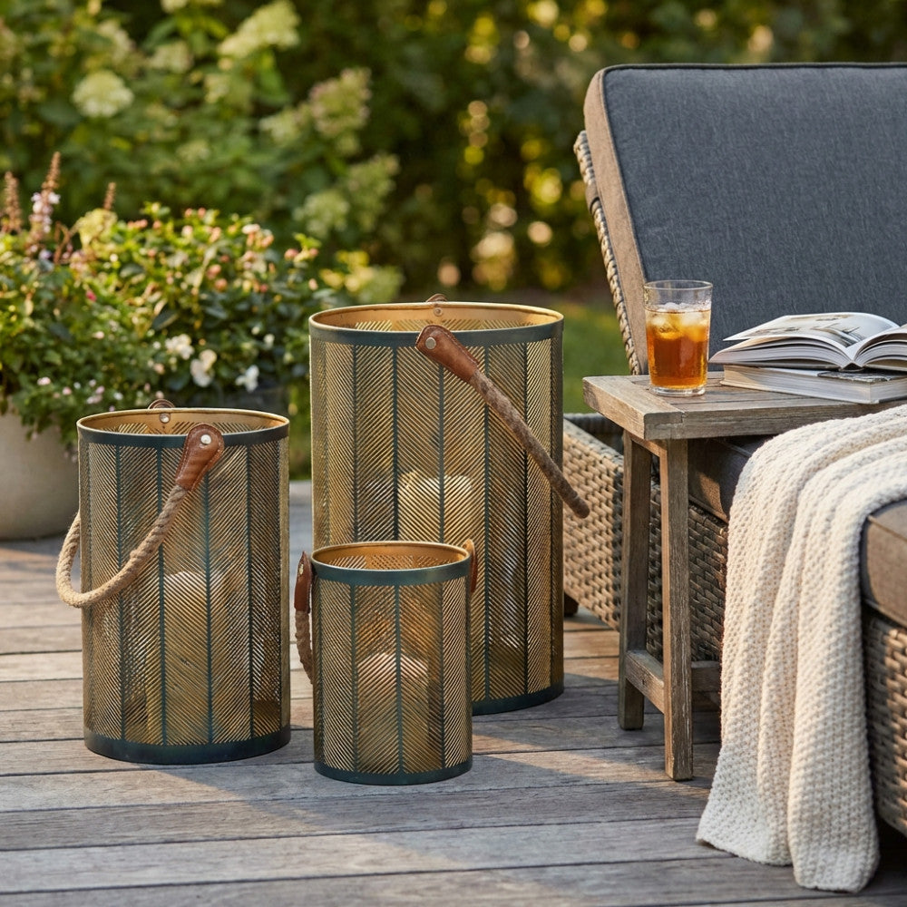 Set di 3 lanterne Chloé in acciaio verde oro con manici in corda, disposte su un patio in legno accanto a una seduta relax.