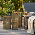 Set di 3 lanterne Chloé in acciaio verde oro con manici in corda, disposte su un patio in legno accanto a una seduta relax.