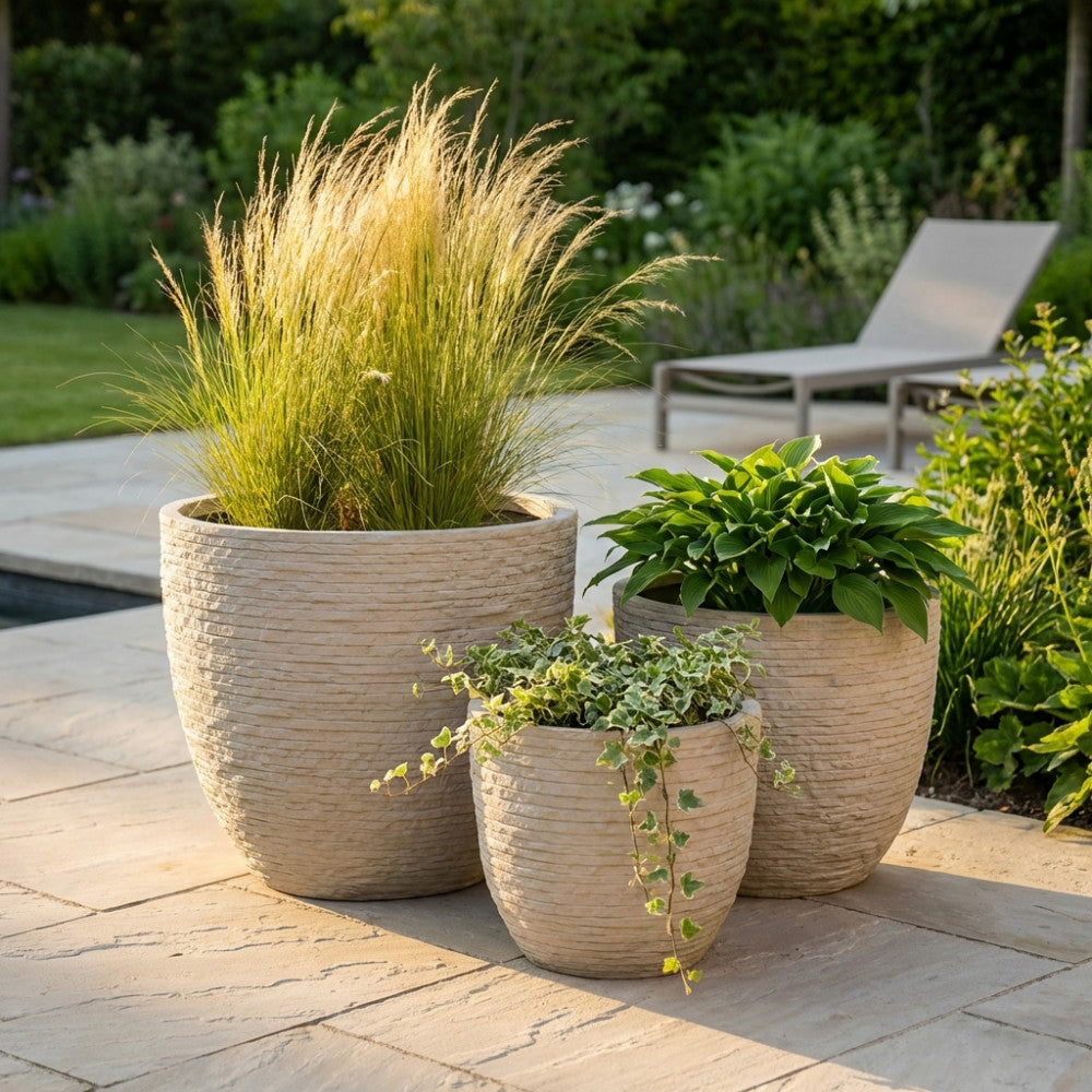 Set di tre vasi Ciclamino in fibra di argilla beige con texture a righe orizzontali, disposti su una pavimentazione in pietra illuminata dal sole.