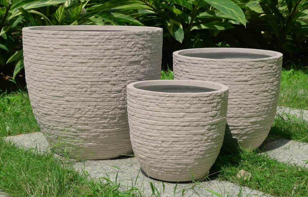 Composizione di tre vasi beige di diverse dimensioni immersi nel verde di un giardino, mostrando l'armonia dei volumi e la resistenza del materiale.
