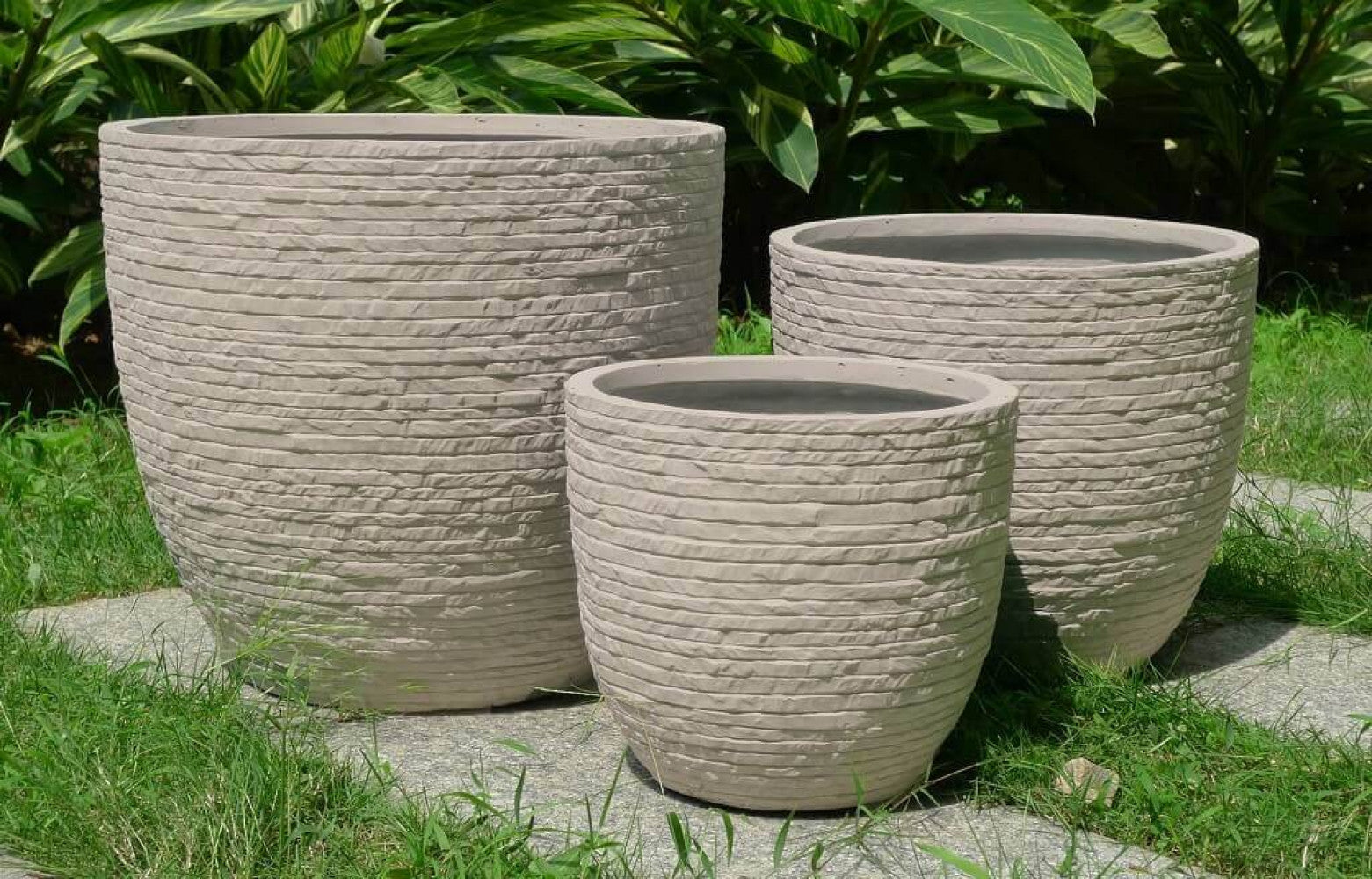 Composizione di tre vasi beige di diverse dimensioni immersi nel verde di un giardino, mostrando l'armonia dei volumi e la resistenza del materiale.