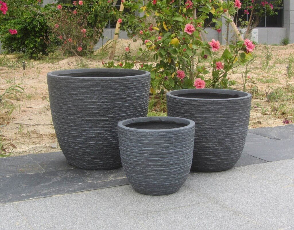 Set di vasi grigi in fibra di argilla posizionati all'aperto su lastre di ardesia, con uno sfondo di arbusti verdi e fiori di ibisco rosa.