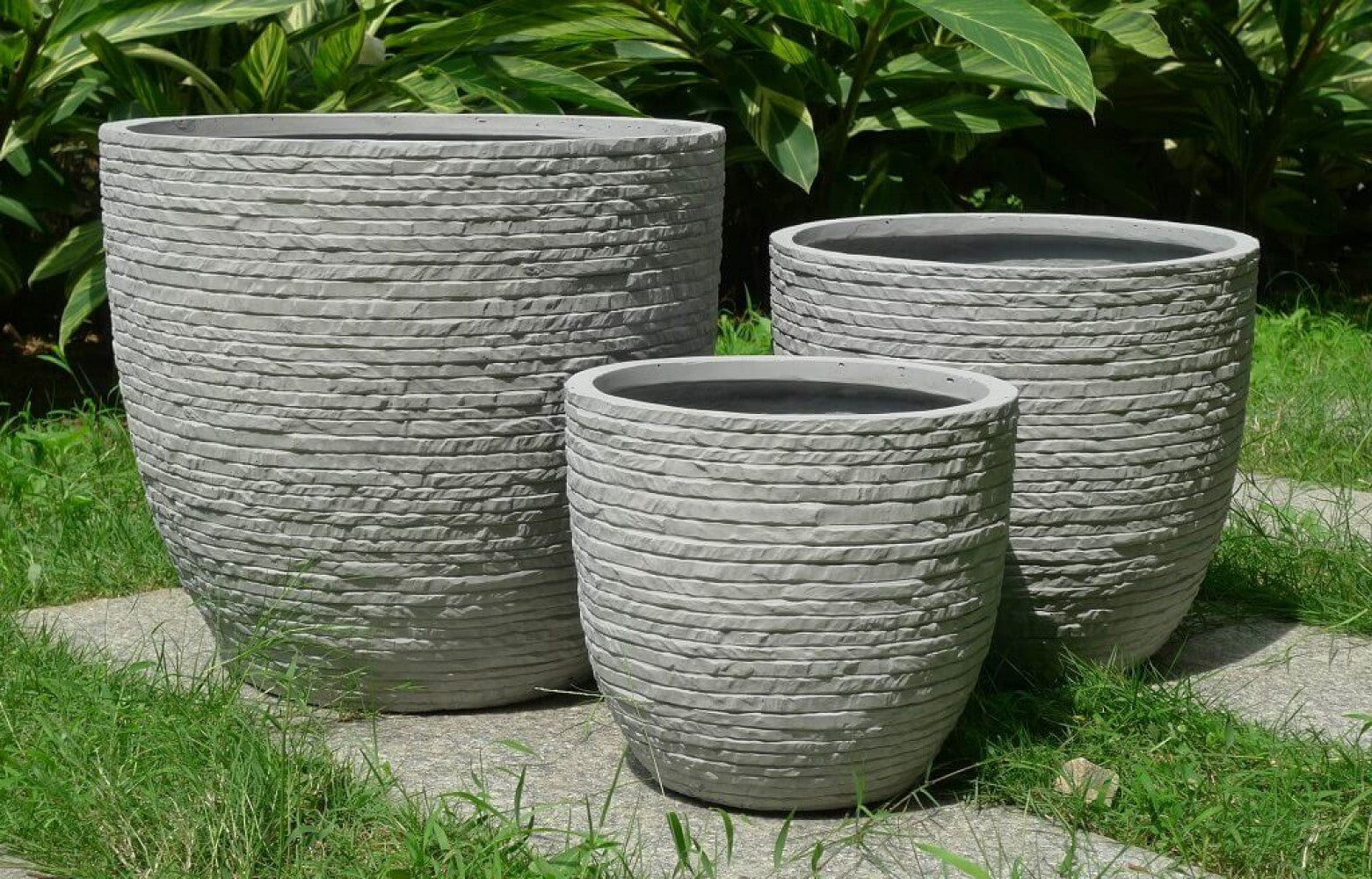 Set di tre vasi decorativi grigi disposti all'aperto su un camminamento in pietra, circondati da erba verde e rigoglioso fogliame tropicale.
