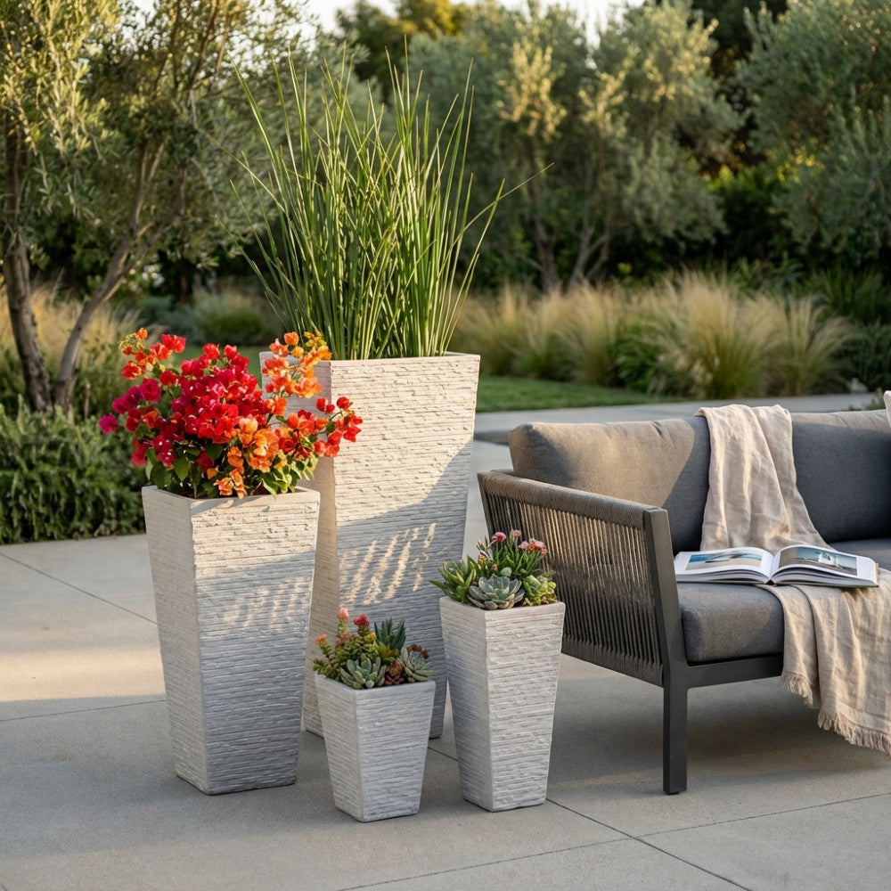 Composizione di vasi scultorei beige di diverse altezze con fiori colorati e piante grasse, posizionati in un giardino lussureggiante al tramonto.