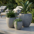 Set di tre vasi Ginepro in fibra sintetica grigia con piante decorative su un patio moderno.