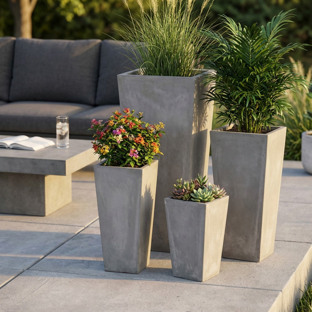 Set di quattro vasi Margherita in fibra di argilla beige disposti su un patio moderno, arricchiti con piante verdi e fiori colorati accanto a un divano grigio.