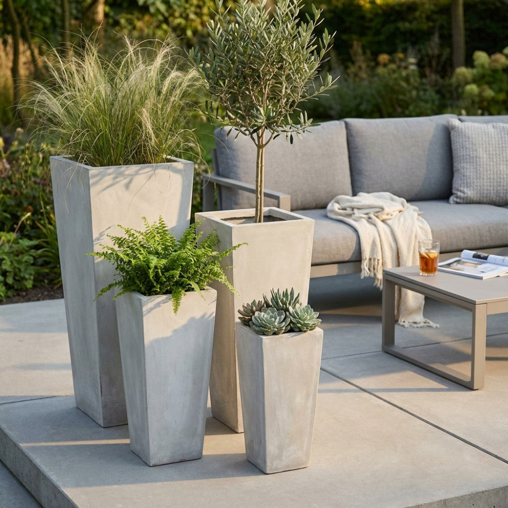 Composizione di vasi geometrici a colonna in fibra di argilla beige su una terrazza elegante, contenenti felci, piante grasse e un piccolo ulivo decorativo.