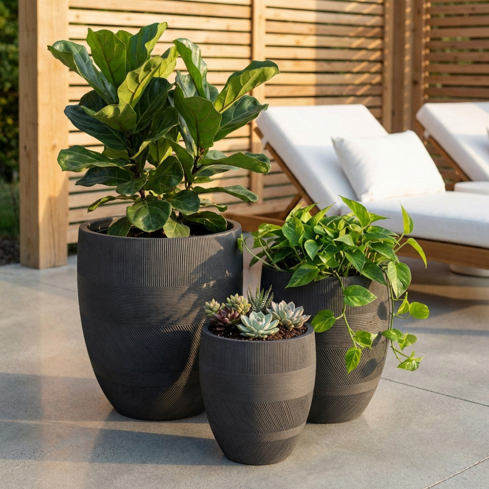 Set di tre vasi in fibra sintetica grigio scuro con texture scanalata, allestiti su un patio moderno con piante verdi e sedute bianche sullo sfondo.