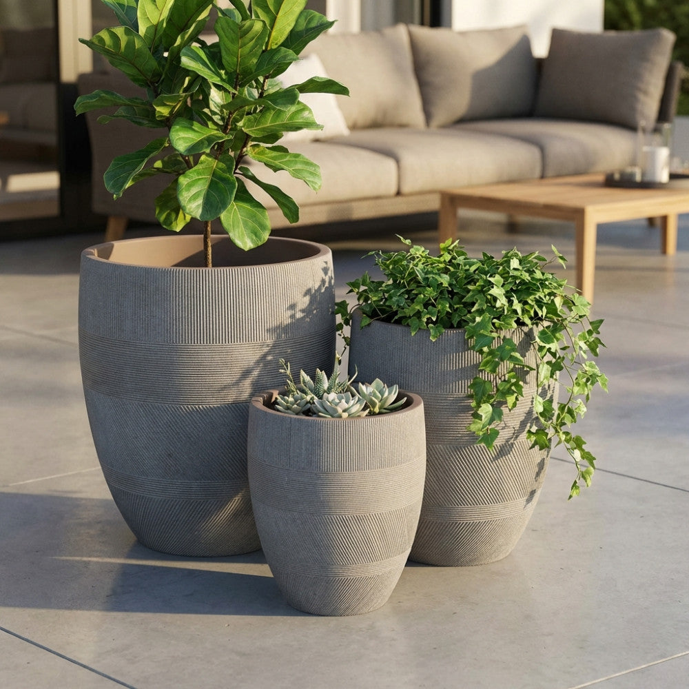Set di tre vasi Papavero color taupe in fibra sintetica disposti su un patio esterno, contenenti piante verdi decorative.