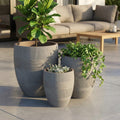 Set di tre vasi Papavero color taupe in fibra sintetica disposti su un patio esterno, contenenti piante verdi decorative.