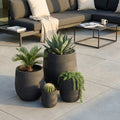Set di quattro vasi Peonia in fibra sintetica grigio scuro, disposti su una terrazza esterna con piante grasse e un divano moderno sullo sfondo.