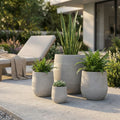 Set di quattro vasi Peonia in fibra sintetica taupe disposti su un patio moderno, arricchiti da piante verdi decorative con un lettino prendisole sullo sfondo.