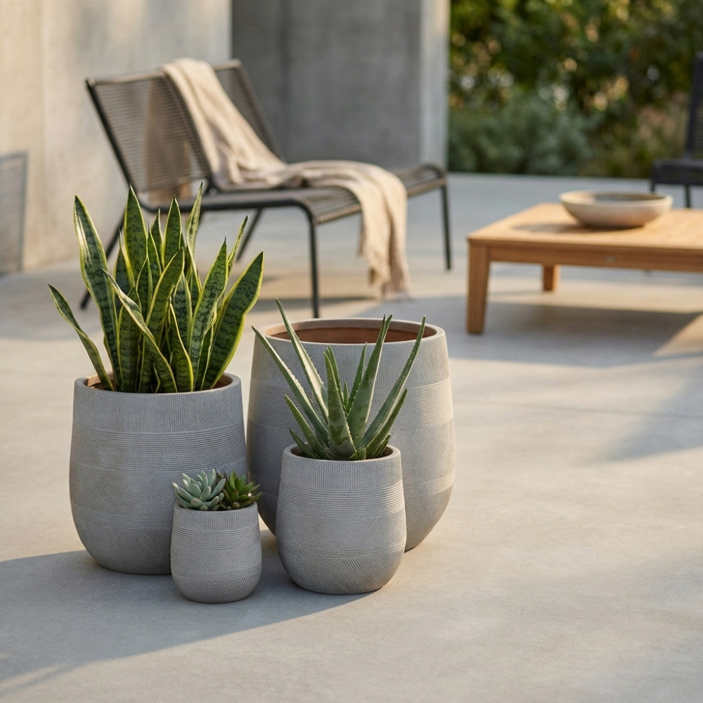 Composizione outdoor con vasi Peonia color taupe di diverse dimensioni, contenenti aloe e sansevieria, affiancati da una sedia di design e un tavolino in legno.