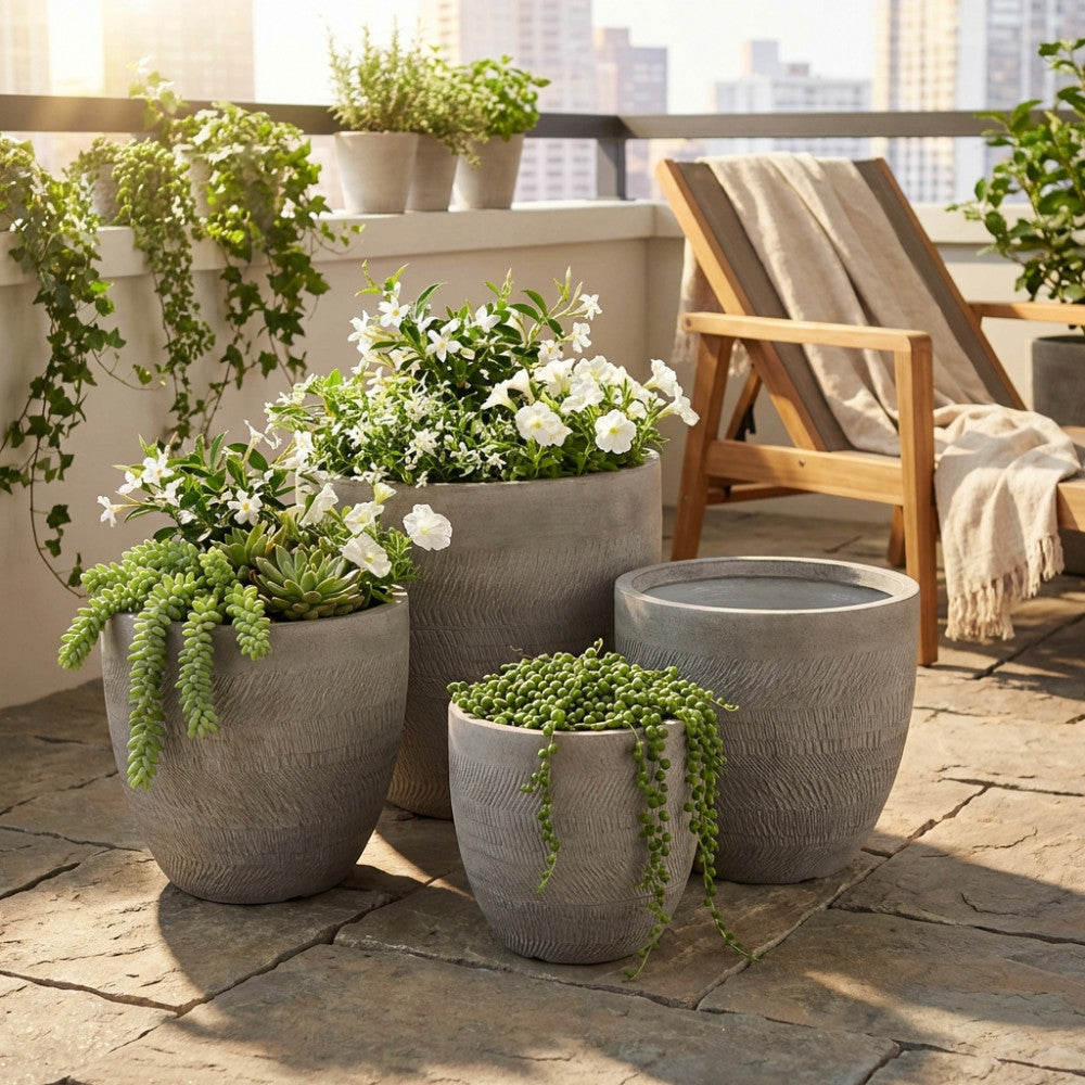 Set di quattro vasi Primula in fibra sintetica grigia, disposti su un patio soleggiato con piante fiorite e succulente.