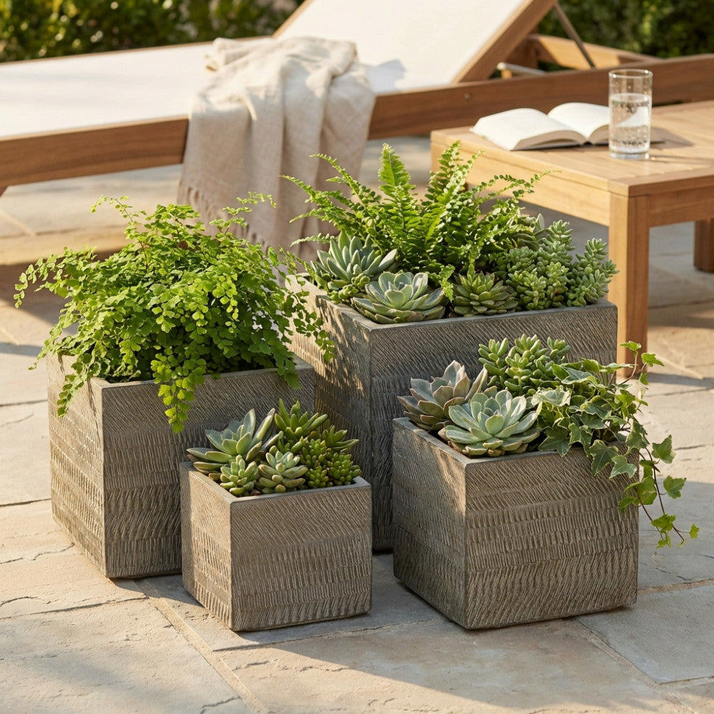 Set di quattro vasi quadrati marroni in fibra sintetica con texture scanalata, allestiti su un patio in pietra con piante grasse e felci.