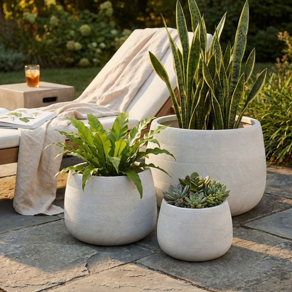 Set di tre vasi Tulipano in fibra sintetica beige posizionati su un patio in pietra, affiancati a un lettino in legno con cuscini chiari e piante ornamentali.