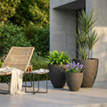 Tre vasi Verbena in fibra sintetica antracite con pattern geometrico, allestiti con piante verdi su un patio moderno accanto a una poltrona.