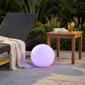 Sfera luminosa Gemini accesa con luce viola, posizionata all'aperto accanto a un lettino e un tavolino in legno.