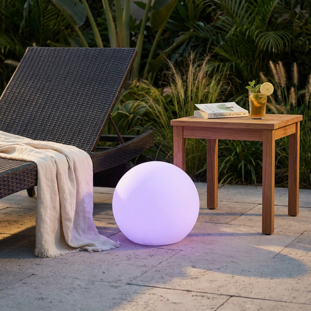Sfera luminosa Gemini accesa con luce viola, posizionata all'aperto accanto a un lettino e un tavolino in legno.
