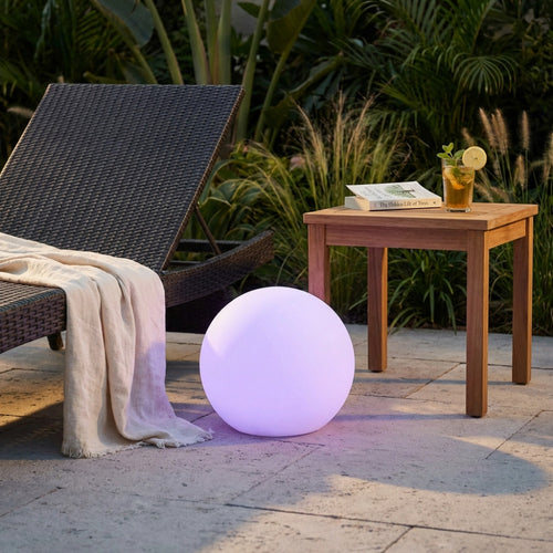 Sfera luminosa Gemini accesa con luce viola, posizionata all'aperto accanto a un lettino e un tavolino in legno.