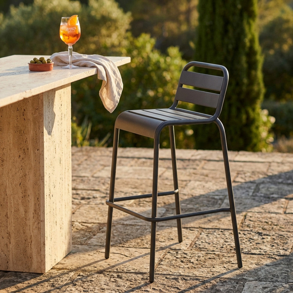 Sgabello alto Vega in acciaio taupe posizionato all'aperto accanto a un bancone in travertino con un cocktail.