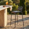 Sgabello alto Vega in acciaio taupe posizionato all'aperto accanto a un bancone in travertino con un cocktail.