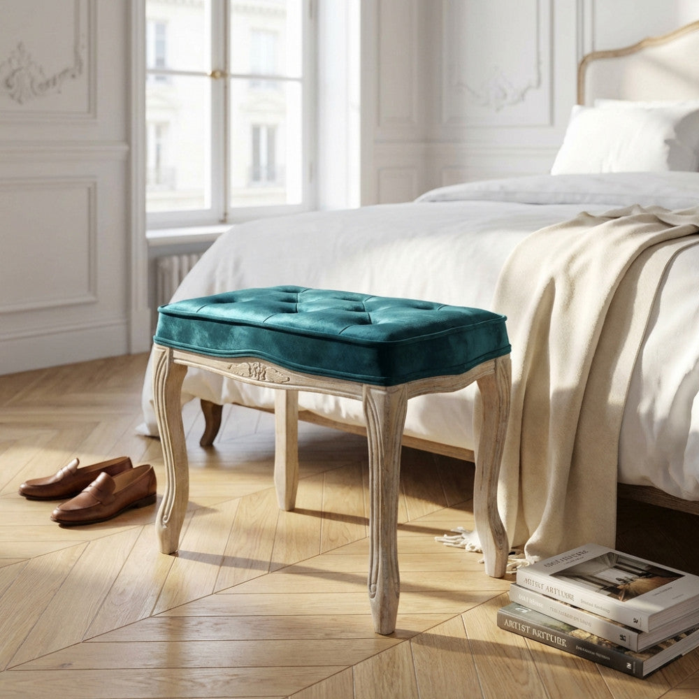 Sgabello Marly in legno e velluto blu posizionato ai piedi di un letto in una camera luminosa.