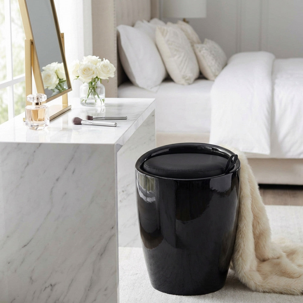 Sgabello pouf Cliff nero lucido posizionato accanto a una toeletta in marmo bianco in una camera da letto luminosa.