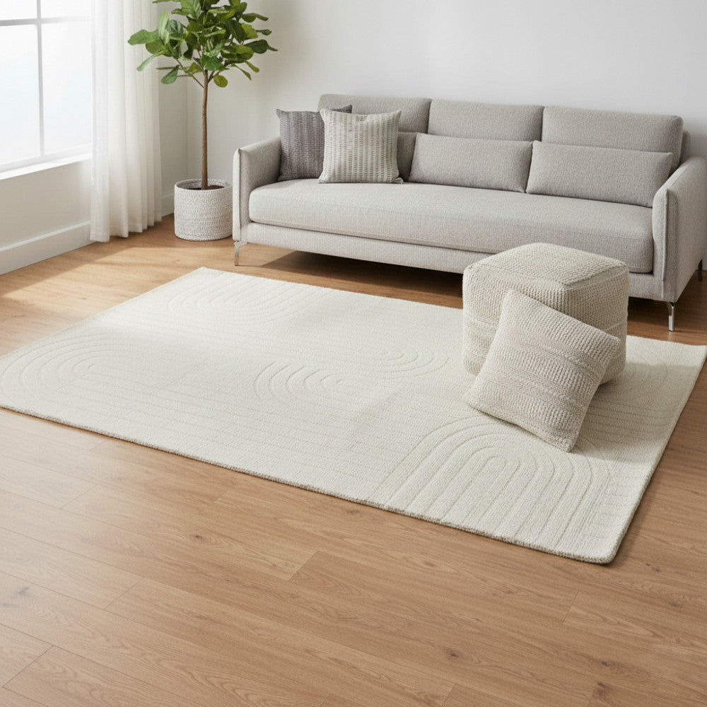 Tappeto Aurélien bianco e beige in un ampio soggiorno minimalista con pouf coordinato.