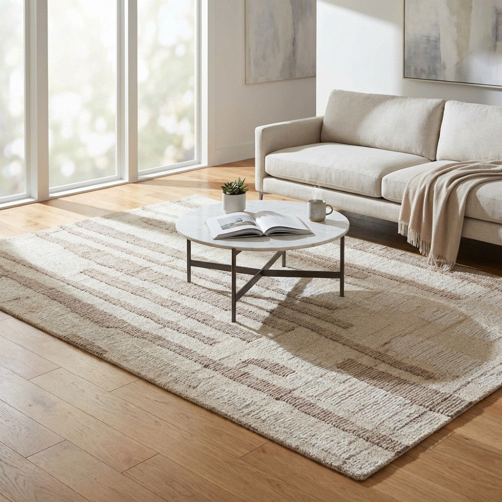 Tappeto moderno Fabien in lana e cotone beige ecrù, posizionato in un luminoso soggiorno con divano bianco e tavolino in marmo.