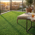Balcone panoramico al tramonto con tappeto erboso sintetico verde, lettino relax intrecciato e tavolino in metallo.