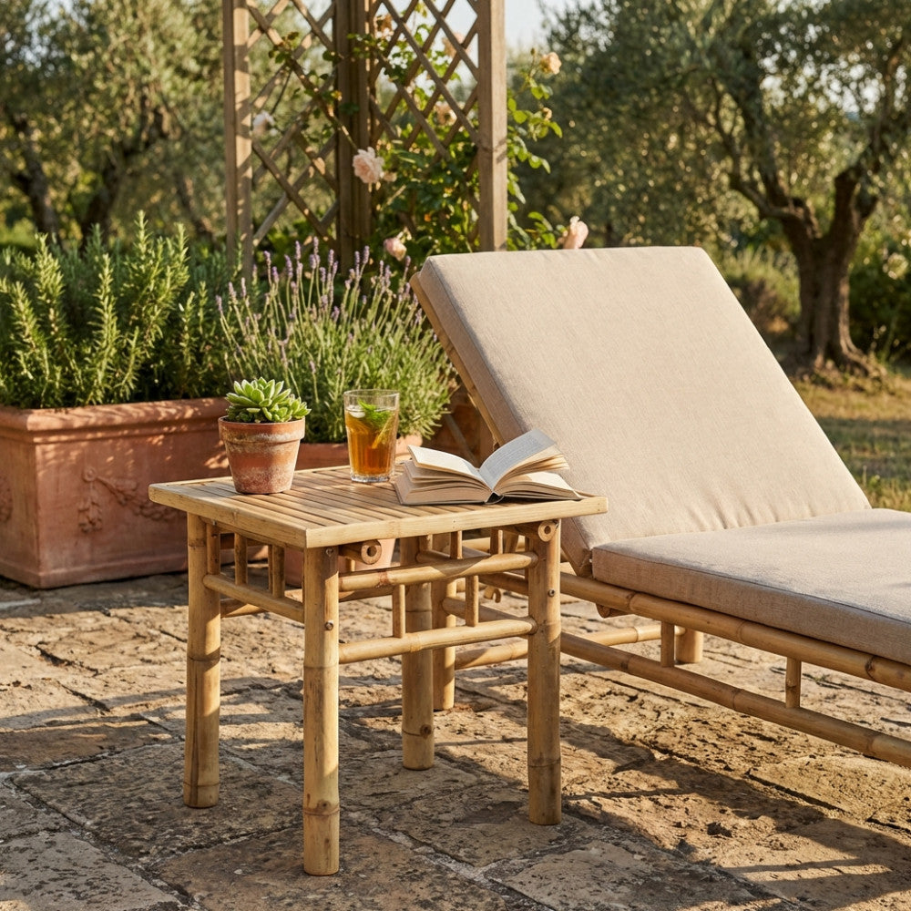 Tavolino da giardino in legno marrone posizionato su un patio in pietra accanto a un lettino prendisole con cuscini chiari.