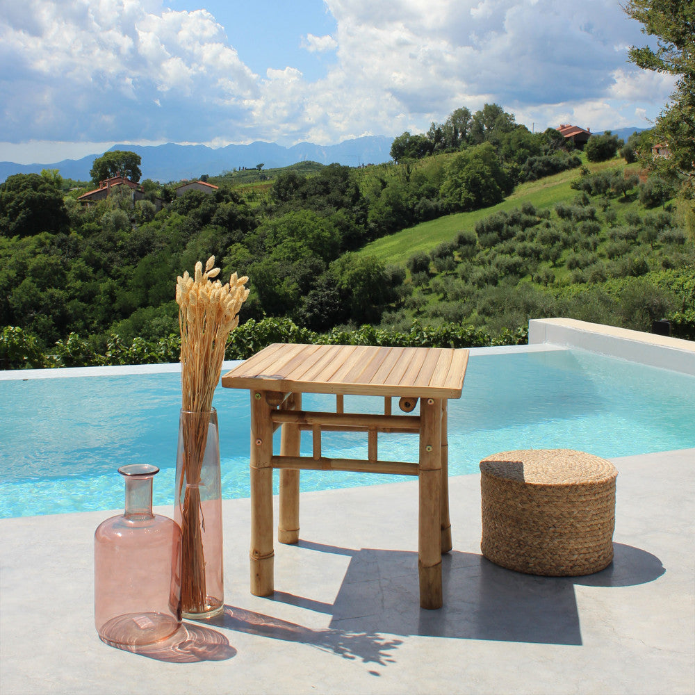 Tavolino in legno marrone allestito a bordo piscina con vasi decorativi in vetro e un pouf in iuta, con vista sulle colline.