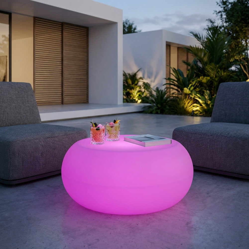 Tavolino luminoso Dorado rosa posizionato all'aperto tra due poltrone grigie, decorato con cocktail e un libro.