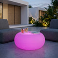 Tavolino luminoso rotondo rosa acceso posizionato su un patio moderno tra due poltrone grigie.
