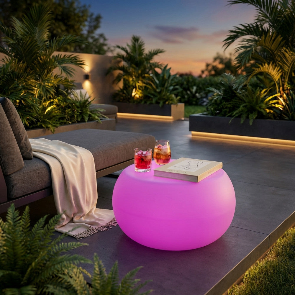 Tavolino luminoso rosa in un giardino al tramonto, accanto a un lettino grigio con drink e un libro sul piano.