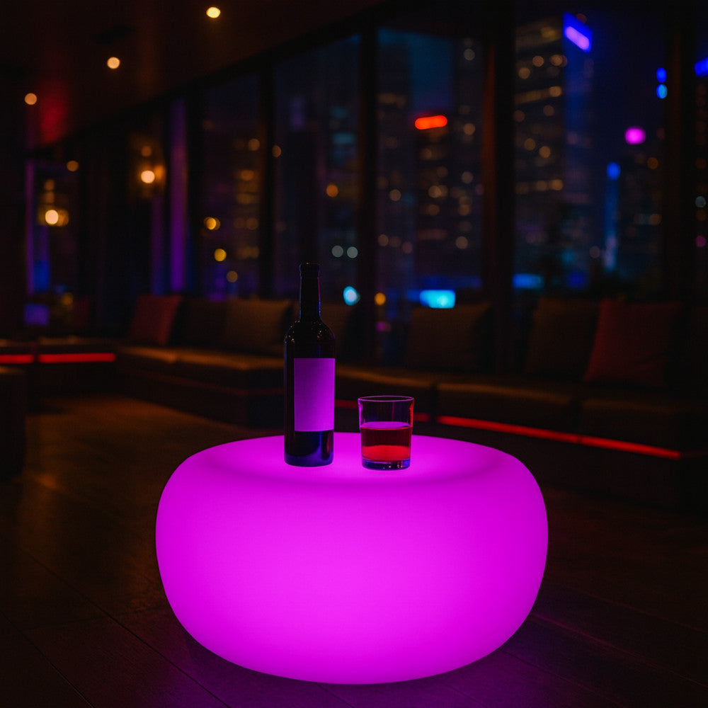 Tavolino luminoso rosa in un ambiente interno notturno con vista sulla città, allestito con bottiglia e bicchiere.
