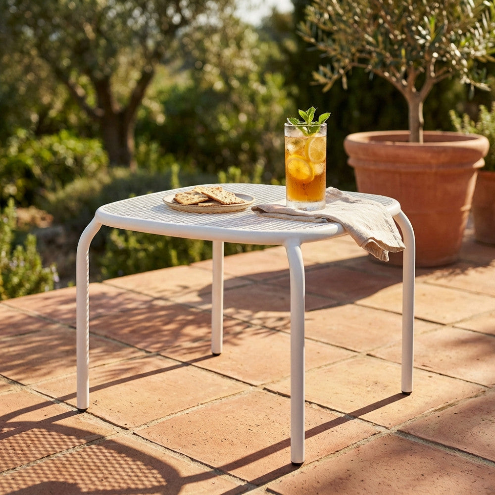 Dettaglio del tavolino da esterno Midway in acciaio bianco, illuminato dalla luce solare su una terrazza rustica con elementi botanici.