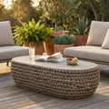 Tavolino ovale Lafayette in corda intrecciata e alluminio, allestito su un patio in legno con libri e frutta, illuminato dalla luce del tramonto.