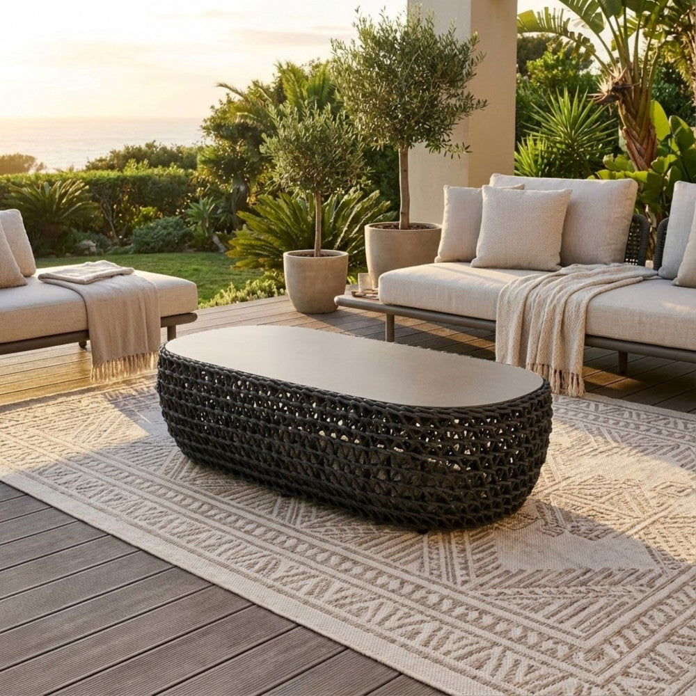Ambiente costiero al tramonto con il tavolino ovale Lafayette su un tappeto geometrico da esterno, circondato da divani lounge e ulivi in vaso.